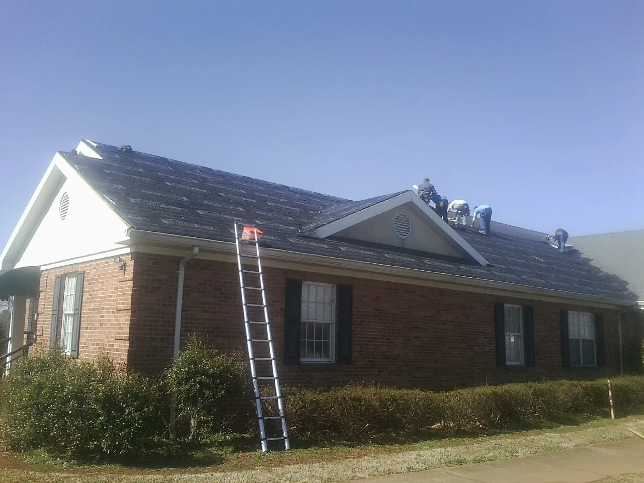Crew tearing shingles off a brick ranch roof with a ladder set — Emergency Roof Repair work by our crew in Manilla